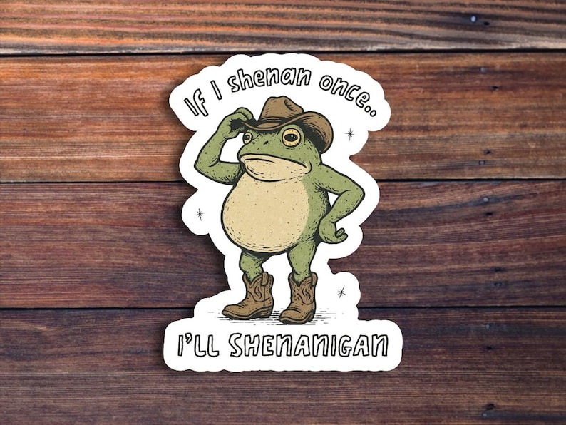 May include: A sticker featuring a cartoon frog wearing a cowboy hat and boots. The frog is green and tan, with the text "If I shenan once... I'll shenanigan" in a playful font. The sticker is on a wooden background.