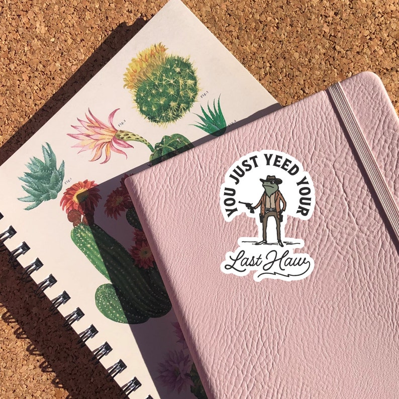 May include: A pink notebook with a sticker on the front. The sticker features a cartoon frog wearing a cowboy hat and holding a gun. The text on the sticker reads "You just yeed your last haw".