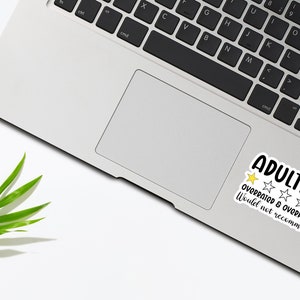 May include: A laptop with a sticker that reads "ADULTING" in black and white, with a yellow star and four white stars. The text "OVERRATED & OVERPRICED. Would not recommend." is also on the sticker. A green leaf is in the lower left corner.