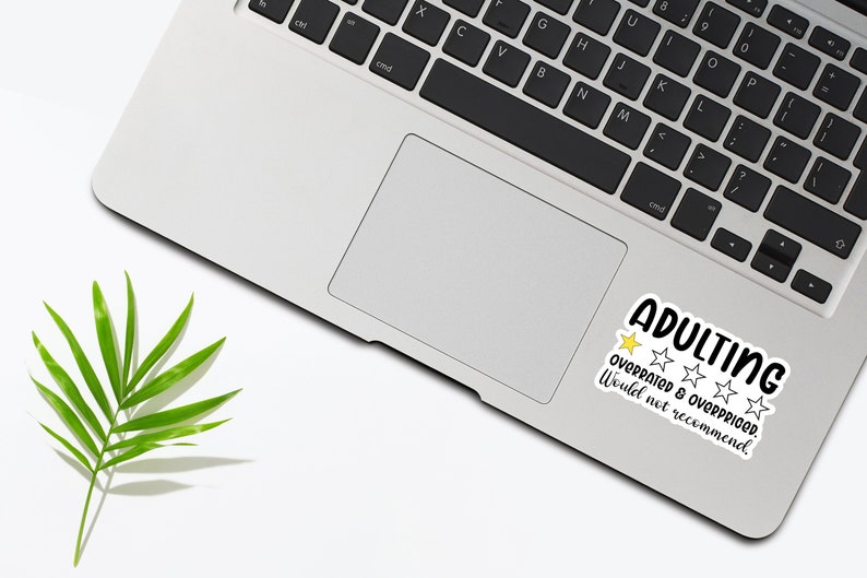 May include: A laptop with a sticker that reads "ADULTING" in black and white, with a yellow star and four white stars. The text "OVERRATED & OVERPRICED. Would not recommend." is also on the sticker. A green leaf is in the lower left corner.