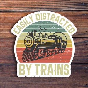 May include: A sticker with a vintage train illustration. The design features a steam engine in black, yellow, and red, set against a striped background of orange, green, and yellow. The text reads "EASILY DISTRACTED BY TRAINS."