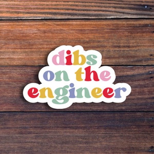 May include: A sticker with the text "dibs on the engineer" in a rainbow of colors. The sticker is on a brown wooden background.