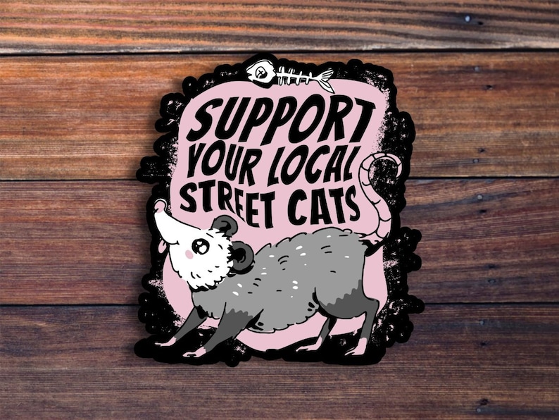 May include: A pink and black sticker with a cartoon opossum and the text "Support your local street cats".