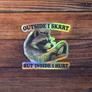 May include: A sticker with a raccoon driving a car. The sticker has a holographic finish and says "Outside I Skrrt But Inside I Hurt."