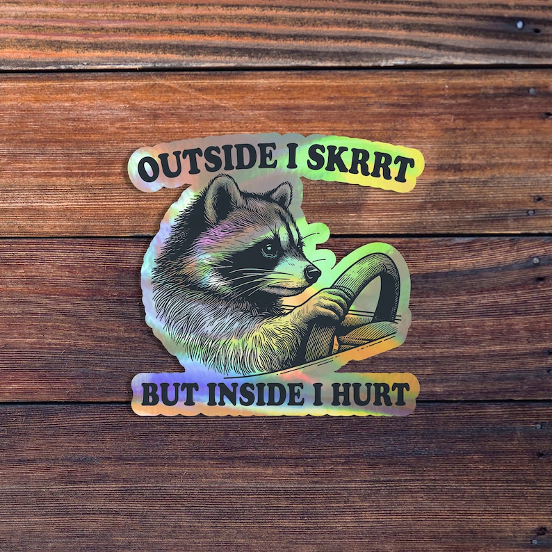 May include: A sticker with a raccoon driving a car. The sticker has a holographic finish and says "Outside I Skrrt But Inside I Hurt."