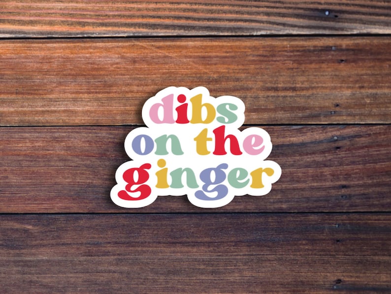 May include: A colorful sticker with the text "dibs on the ginger" in a retro font style. The sticker is on a brown wooden background.