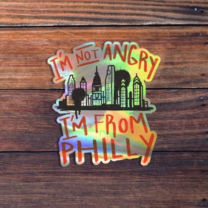May include: A holographic sticker with the text "I'm not angry I'm from Philly" and a black and white silhouette of the Philadelphia skyline.