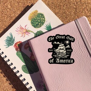 May include: A black and white sticker with the text "The Great Gulf of America" in a gothic font. The sticker features a ship sailing on the sea with clouds and a sun in the sky. The text "Est. 2025" is below the ship.