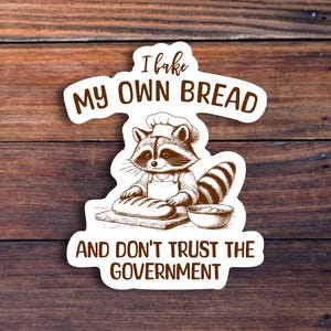 May include: A sticker featuring a raccoon chef baking bread, with the text "I bake my own bread and don't trust the government." The raccoon is wearing a chef's hat and apron, with a loaf of bread on a cutting board.