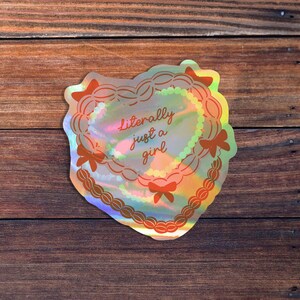 May include: A holographic sticker with a heart-shaped design and the text "literally just a girl". The sticker is iridescent and has a pink and blue color scheme.