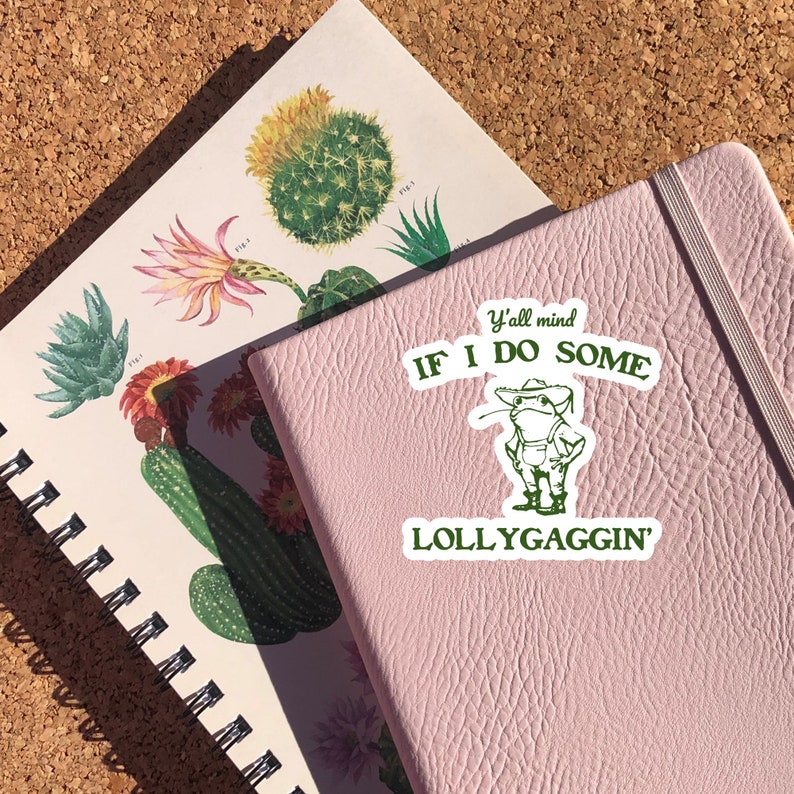 May include: A pink notebook with a green sticker that says "Y'all mind if I do some lollygaggin'" with a cartoon frog wearing a cowboy hat and overalls.