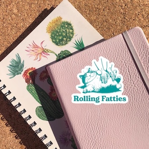 May include: A teal green sticker with a cartoon illustration of a cat being petted by two hands. The text "Rolling Fatties" is printed below the cat.