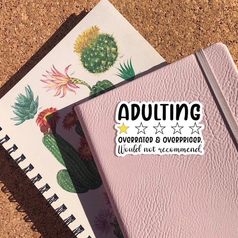 May include: A sticker with the word "ADULTING" in bold black letters, with a rating of one gold star and four white stars, and the text "overrated & overpriced. Would not recommend." The sticker is on a pink notebook next to a botanical print notebook.