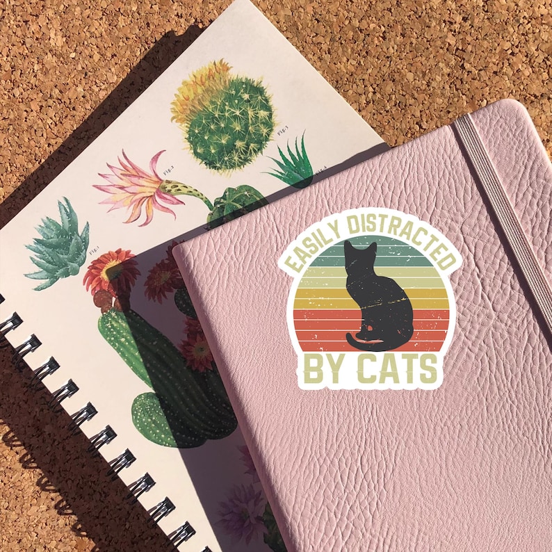 May include: A pink notebook with a sticker that says "Easily Distracted By Cats". The sticker features a black silhouette of a cat sitting in a retro-style sunburst with orange, yellow, and red stripes.