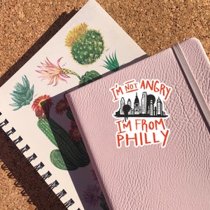 May include: A pink notebook with a sticker that says "I'm not angry I'm from Philly" with a black silhouette of the Philadelphia skyline.