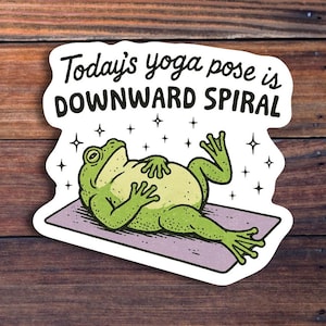 May include: A sticker featuring a cartoon frog in a yoga pose. The frog is green with a yellow belly, lying on a purple mat. The text reads "Today's yoga pose is DOWNWARD SPIRAL" in a playful font, with star accents.