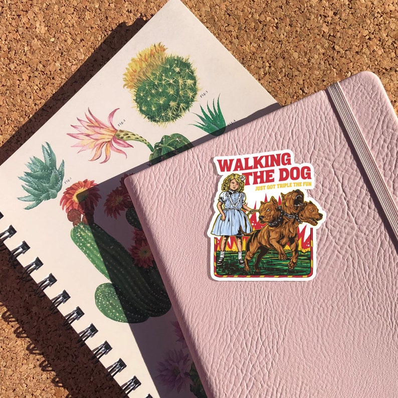 May include: A pink notebook with a sticker on the front. The sticker features a cartoon girl walking a three-headed dog. The sticker says "Walking the Dog. Just got triple the fun."