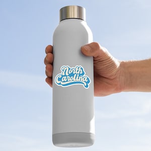 K&ouml;nnte beinhalten: Eine graue Wasserflasche mit einem blauen und wei&szlig;en Aufkleber, auf dem in einer Retro-Schrift "North Carolina" steht.