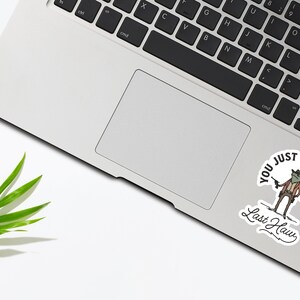May include: A white laptop computer with a sticker on the trackpad. The sticker features a cartoon frog wearing a cowboy hat and holding a gun. The text on the sticker reads "You just yeed your last haw".