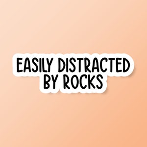 May include: A white sticker with a black border and the text "EASILY DISTRACTED BY ROCKS" in a bold, black font. The sticker is set against a peach-colored background.