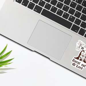 May include: A laptop with a keyboard and trackpad, with two stickers on the surface. One sticker features a mouse in a cloak holding a sword, with the text "I'm Burnt out M'Lord". A green leaf is on the white surface.