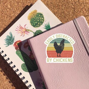 May include: A pink notebook with a sticker that says "Easily Distracted by Chickens". The sticker features a black rooster with a red, orange, and yellow sunset background.