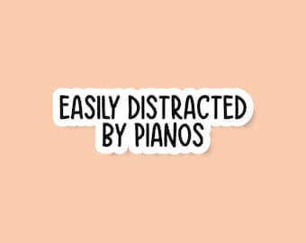 May include: A white sticker with black text that reads "Easily Distracted by Pianos" on a peach background.