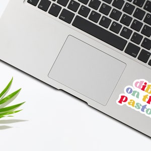 K&ouml;nnte beinhalten: Ein Laptop mit einem Aufkleber, auf dem "dibs on the pastor" in Regenbogenfarben steht. Der Aufkleber befindet sich auf dem Trackpad des Laptops.