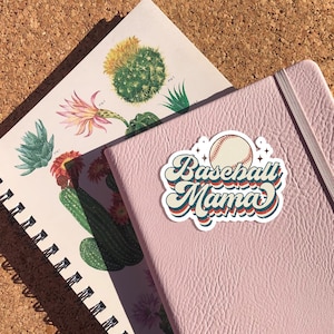 Pode incluir: Um caderno rosa com um adesivo branco que diz "Baseball Mama" em uma fonte retr&ocirc;. O adesivo tamb&eacute;m apresenta uma bola de beisebol.
