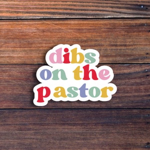K&ouml;nnte beinhalten: Ein bunter Aufkleber mit dem Text "dibs on the pastor" in einem Regenbogen-Farbschema. Der Aufkleber befindet sich auf einem braunen Holzhintergrund.