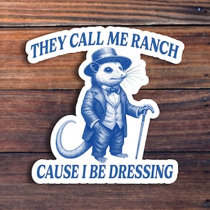 May include: A blue and white sticker featuring a dapper opossum in a suit, hat, and cane. The text reads "THEY CALL ME RANCH CAUSE I BE DRESSING". The sticker is set against a wooden background.