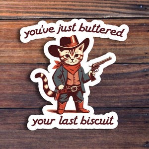 May include: A white sticker with a cartoon cat wearing a cowboy hat and holding a gun. The text on the sticker reads "You've just buttered your last biscuit."