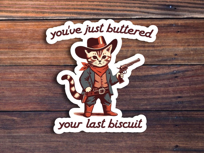 May include: A white sticker with a cartoon cat wearing a cowboy hat and holding a gun. The text on the sticker reads "You've just buttered your last biscuit."
