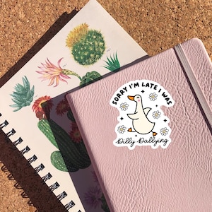 May include: A pink notebook with a white sticker featuring a cartoon duck with the text "Sorry I'm late I was dilly dallying" and white flowers.