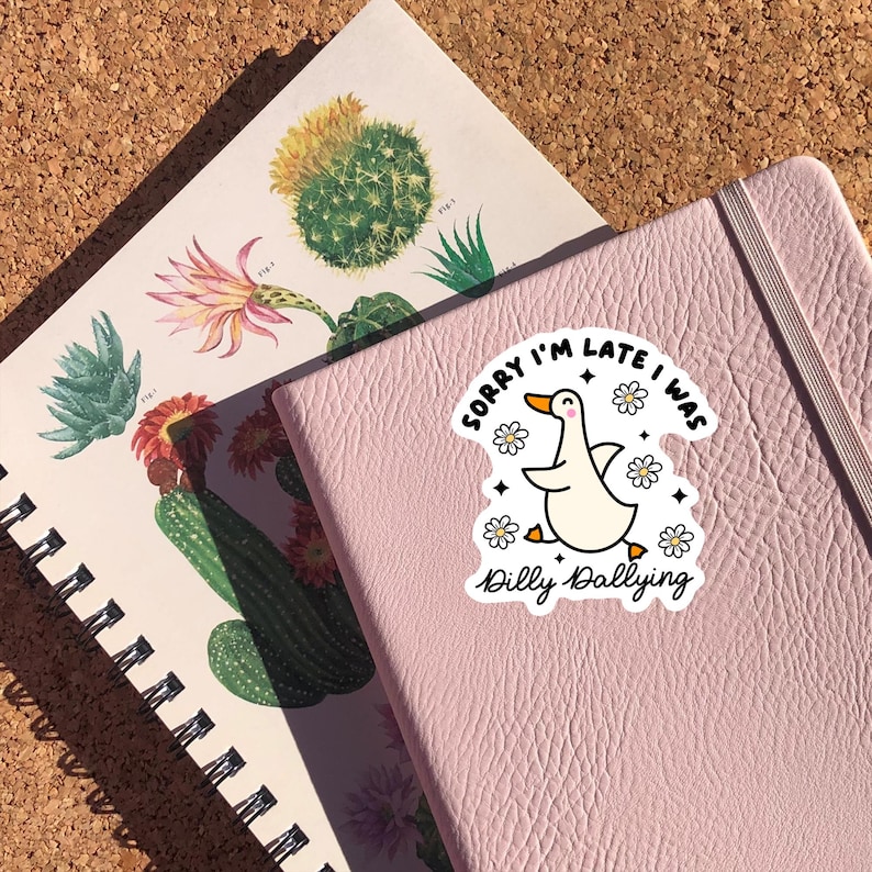 May include: A pink notebook with a white sticker featuring a cartoon duck with the text "Sorry I'm late I was dilly dallying" and white flowers.