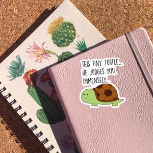 May include: A pink notebook with a sticker of a green turtle with a brown shell. The sticker says "This tiny turtle... he judges you immensely..."