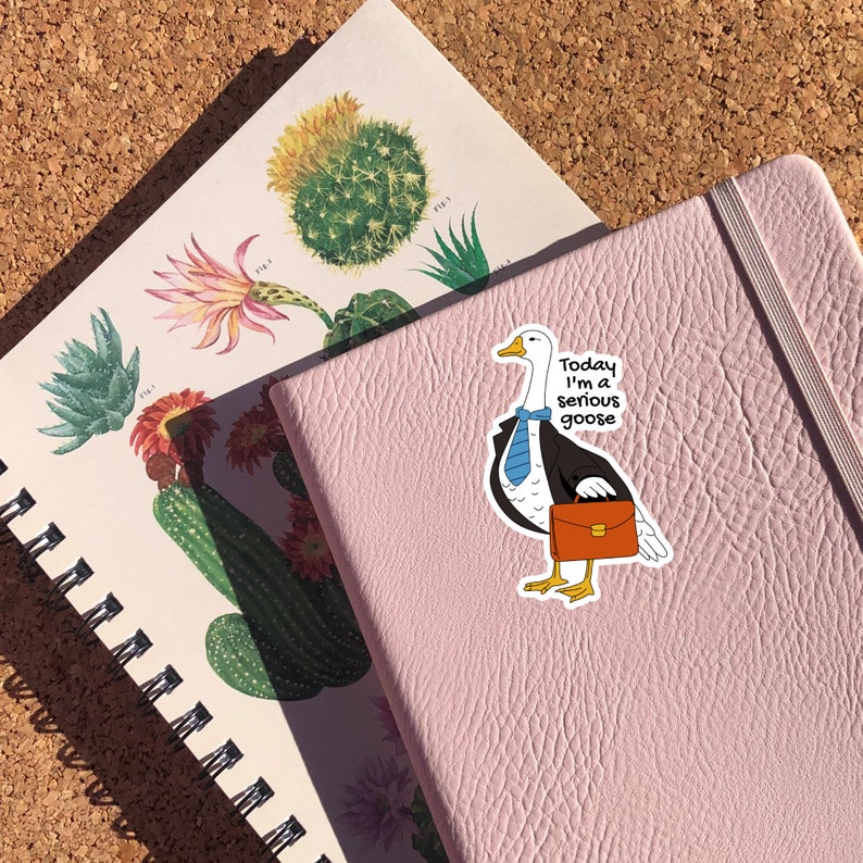May include: A pink notebook with a white goose sticker on it. The goose is wearing a blue tie and a black suit and holding a brown briefcase. The sticker says "Today I'm a serious goose."