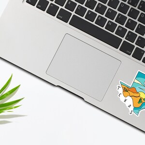 May include: A sticker of the state of Texas with a colorful landscape design. The sticker is on a silver laptop.