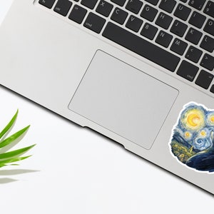 K&ouml;nnte beinhalten: Ein Laptop mit einem Aufkleber eines schwarzen Katers, der zu einem sternenklaren Nachthimmel aufblickt. Der Aufkleber ist eine Nachbildung des ber&uuml;hmten Gem&auml;ldes "Sternennacht" von Vincent van Gogh.
