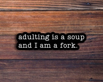 Op de afbeelding: Een zwart-witte sticker met de tekst "adulting is a soup and I am a fork." op een bruine houten achtergrond.
