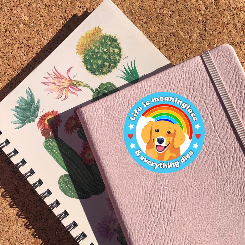 Puede incluir: Una libreta rosa con una pegatina azul y blanca que dice "La vida no tiene sentido y todo muere". La pegatina presenta un golden retriever de dibujos animados con un arco&iacute;ris detr&aacute;s.