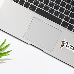 May include: Laptop with a sticker featuring medieval illustrations and the text "That was a bit cringe, my liege." A green leaf is on the white surface.