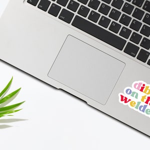 May include: A laptop computer with a sticker that says "dibs on the welder" in rainbow colors. The sticker is on the trackpad of the laptop.