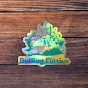May include: A holographic sticker with a green cartoon cat being rubbed on the belly by two hands. The text "Rolling Fatties" is printed below the cat.
