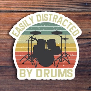 May include: A vintage-style sticker with the text "Easily Distracted By Drums" in a white circle with a red, orange, and yellow striped background. A black silhouette of a drum set is in the center of the circle.