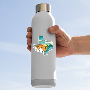 May include: A stainless steel water bottle with a sticker of the state of Texas. The sticker features a colorful illustration of the state with a sunset, mountains, and a river. The text "TEXAS" is written in white letters on the sticker.