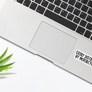 Op de afbeelding: Een witte laptop met een zwart toetsenbord en een sticker met de tekst "Gemakkelijk afgeleid door motorfietsen".