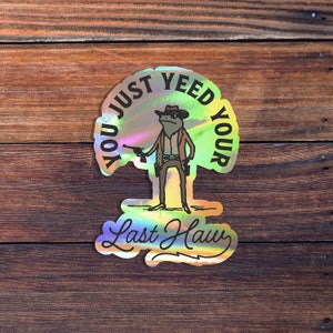 May include: A holographic sticker with a cartoon frog wearing a cowboy hat and holding a gun. The text on the sticker reads "You Just Yee'd Your Last Haw".