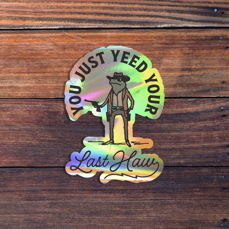 May include: A holographic sticker with a cartoon frog wearing a cowboy hat and holding a gun. The text on the sticker reads "You Just Yee'd Your Last Haw".