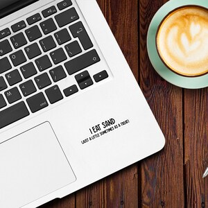 May include: A white laptop computer with a black keyboard and a black trackpad. A sticker on the laptop reads "I eat sand (just a little sometimes as a treat)".
